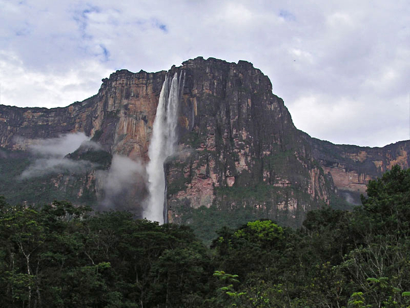 Montanha mostrada mais ao fundo da imagem, com a floresta aparecendo em primeiro plano. No centro da montanha tem uma grande queda de água.